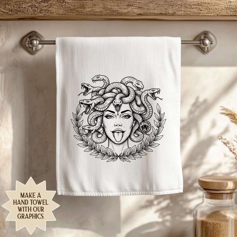 May include: White hand towel featuring a black and white illustration of Medusa with snakes for hair, surrounded by a laurel wreath. The towel is hanging on a metal bar. Text on the bottom left says "MAKE A HAND TOWEL WITH OUR GRAPHICS."