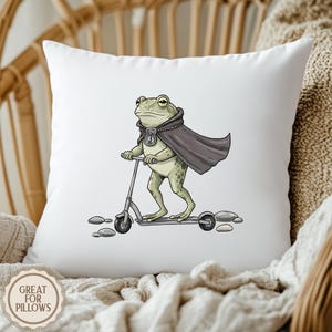 K&ouml;nnte beinhalten: Wei&szlig;es Kissen mit einem Cartoon-Frosch auf einem Roller. Der Frosch tr&auml;gt einen dunkelgrauen Umhang und eine Halskette. Das Kissen hat den Text "GREAT FOR PILLOWS" in einem kreisf&ouml;rmigen Design.