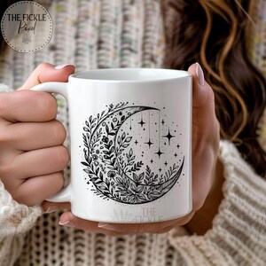 May include: White ceramic mug with a black and white illustration of a crescent moon surrounded by leaves and flowers. The moon has stars hanging from it.