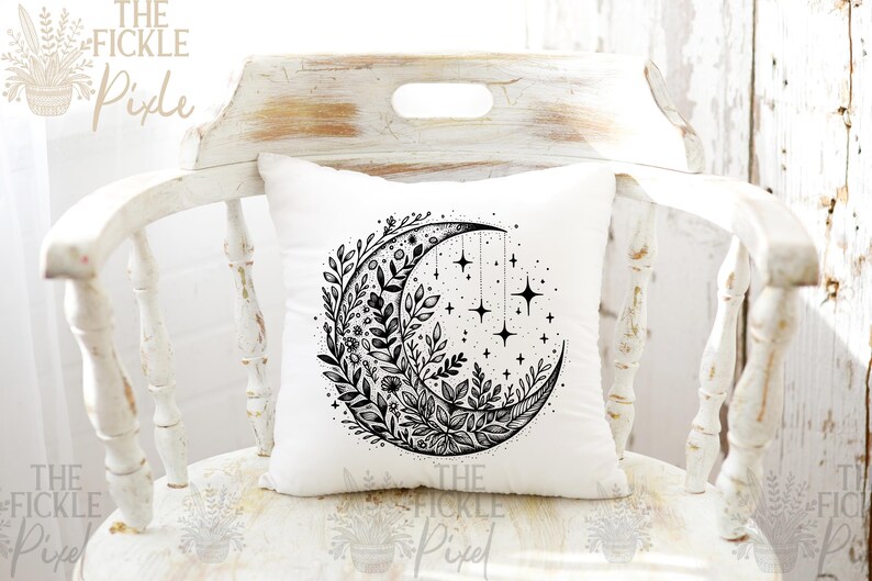 May include: A white pillow with a black and white illustration of a crescent moon with floral designs and stars. The moon is surrounded by leaves and flowers.
