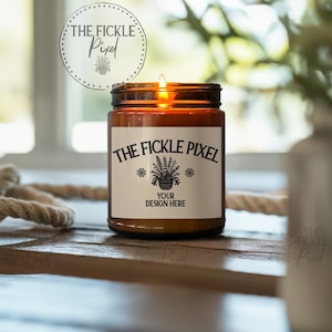May include: A lit candle in a brown glass jar with a white label. The label reads "THE FICKLE PIXEL" above a plant illustration and "YOUR DESIGN HERE". The candle sits on a wooden surface with a rope and greenery in the background.