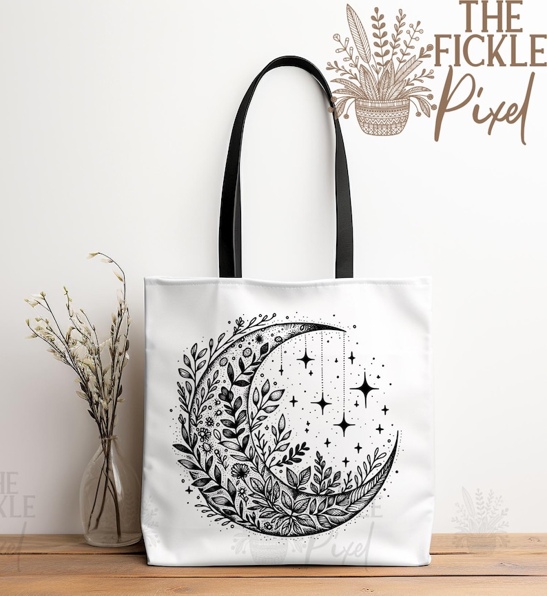 May include: A white tote bag with black handles features a black and white illustration of a crescent moon surrounded by floral designs and stars. The text "The Fickle Pixel" is printed in brown on the top right corner of the bag.