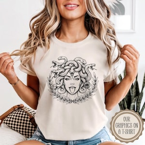 May include: A beige t-shirt featuring a black and white graphic of Medusa with snakes for hair, a tongue sticking out, and a laurel wreath. The text "OUR GRAPHICS ON A T-SHIRT" is printed on a circular emblem on the bottom right.
