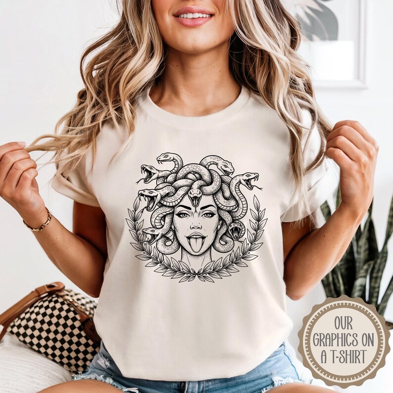 May include: A beige t-shirt featuring a black and white graphic of Medusa with snakes for hair, a tongue sticking out, and a laurel wreath. The text "OUR GRAPHICS ON A T-SHIRT" is printed on a circular emblem on the bottom right.