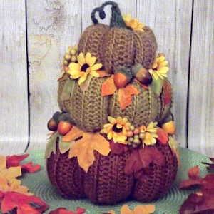May include: A stack of three knitted pumpkins decorated with fall leaves, sunflowers, and acorns. The pumpkins are brown and the leaves are orange, red, and yellow.
