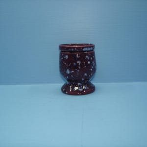 May include: A small, dark maroon ceramic egg cup featuring a glossy finish with a speckled white and light blue pattern throughout the exterior. The cup has a rounded bowl shape, a slightly flared rim, and a sturdy pedestal base.