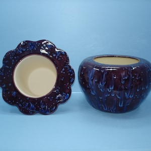 May include: Two ceramic planters. One is a round, dark purple pot with blue drips. The other is a dark purple, flower-shaped planter with a cream-colored interior. Both planters sit on a light blue surface.
