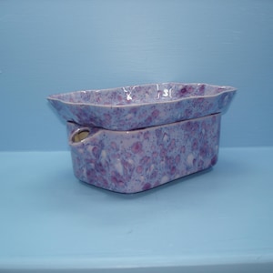 May include: A rectangular ceramic wax warmer with a speckled lavender and purple glaze. The piece consists of a base with a small circular opening on the side and a matching shallow dish top with wavy, uneven edges for holding wax melts.