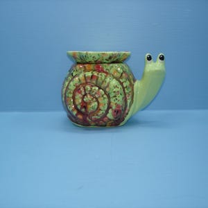 May include: A ceramic snail-shaped planter featuring a light green body and a shell with a mottled glaze of green, orange, and brown tones. The snail has two small eyes on stalks and a wide, circular opening at the top of its shell.