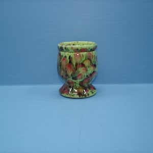 May include: A ceramic egg cup featuring a glossy finish with a mottled pattern of light green, dark green, and reddish-brown spots. The cup has a rounded bowl shape that tapers down to a flared circular base, providing stability.