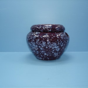 May include: A round, dark purple ceramic yarn bowl with a glossy finish and a white speckled pattern. The bowl features a smooth, rounded rim and a small circular opening on the side for yarn to pass through, creating a functional vessel.