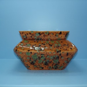 May include: A ceramic, geometric-shaped container with a lid. The container is a warm orange color with a speckled pattern of green and black. The lid is a smaller, rectangular shape that mirrors the container's design. The background is a solid light blue.