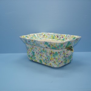May include: A rectangular ceramic soap dish with a wavy rim and a matching base, featuring a speckled pattern of teal, yellow, and purple dots on a white background. The dish has a small spout on one side for drainage and a glossy finish.