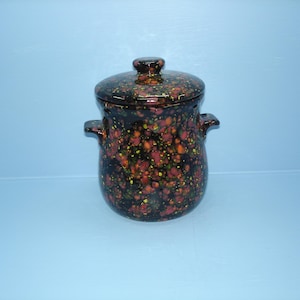 May include: A speckled ceramic pot with a lid. The pot is dark brown with red, orange, and yellow speckles. It has two handles and a matching lid with a small knob. The pot is on a light blue surface.