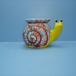 May include: A ceramic snail-shaped wax warmer featuring a bright yellow head and neck. The shell is decorated with a marbled pattern of blue, red, and white glaze, topped with a removable dish for wax melts, creating a functional decorative piece.