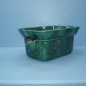 May include: A rectangular ceramic wax warmer with a glossy finish in shades of deep green and brown speckled accents. The piece features a removable top dish with wavy edges and a base with a small circular opening for a heating element.