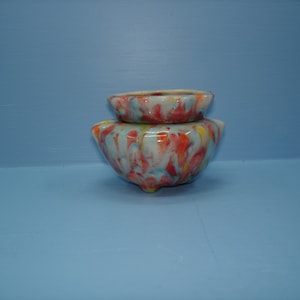 May include: A small, round, footed glass bowl featuring a marbled pattern of white, red, orange, and light blue swirls. The bowl has a wide, open top with a slightly flared rim and a smooth, glossy finish throughout its entire exterior surface.