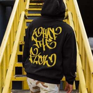 Może przedstawiać: Czarna bluza z kapturem z żółtym napisem w stylu graffiti na plecach o treści "WHAT THE FLOW". Bluza ma długie rękawy i kaptur, noszona na jasnych spodniach moro, podczas stania na żółtych metalowych schodach.