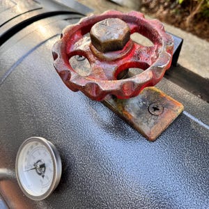 May include: Close-up of a black grill with a red, rusty valve and a temperature gauge. The valve is circular with a hexagonal nut in the center. The temperature gauge has a white face and a black needle.