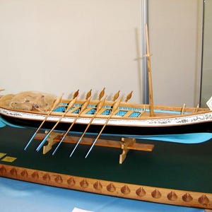 May include: A detailed model of a wooden rowing boat with a blue and black hull, white trim, and a single mast. The boat is displayed on a wooden base with a green felt surface. The model features multiple oars and is a realistic representation of a traditional rowing vessel.