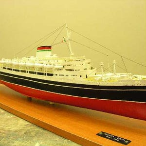 Può includere: Un modello di nave bianco e nero con accenti rossi. La nave ha un ponte bianco e uno scafo nero con una striscia rossa. La nave è su una base di legno con una targa che recita "Andrea Doria - 1953".