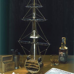 May include: A detailed model of the USS Constitution, a historic sailing ship, with intricate rigging and a blue flag at the masthead. The model is displayed on a wooden surface, alongside a bottle of whiskey, a glass, and a book titled "Old Ironsides".