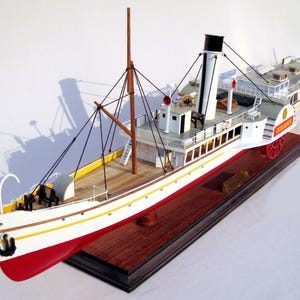 Op de afbeelding: Een gedetailleerd model van een witte en rode stoomboot met een zwarte schoorsteen. Het model staat op een houten basis en heeft een naamplaatje met de tekst "The Islander".