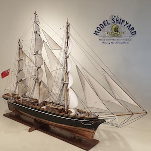 May include: A detailed model of a sailing ship with white sails and a black hull. The ship is on a wooden stand and has a red flag with a white cross on it. The text "The Model Shipyard" is visible in the upper right corner of the image.