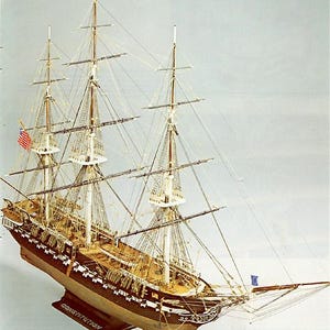 Könnte beinhalten: Ein detailliertes Modell der USS Constitution, ein berühmtes amerikanisches Kriegsschiff, mit aufwendiger Takelage und Segeln. Das Modell ist aus Holz gefertigt und in dunkelbraun lackiert. Der Name des Schiffes, "Constitution", ist auf dem Sockel zu sehen.