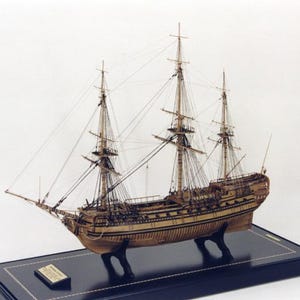 May include: A detailed wooden model of a ship with three masts and a black and white striped hull. The ship is mounted on a black wooden base with a gold trim.