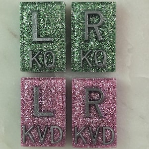 May include: Four square tiles with glitter. Two tiles are green with silver letters 'L' and 'R' and the words 'KQ'. Two tiles are pink with silver letters 'L' and 'R' and the words 'KVD'.