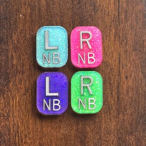 May include: Four rectangular resin tags with rounded corners, featuring glittery finishes in light blue, hot pink, purple, and neon green. Each tag displays silver-coloured embossed lettering with an L or R and the letters NB underneath.