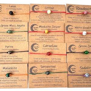 May include: A selection of crystal healing bracelets, each with a different gemstone like lava, citrine, and rhodonite. The bracelets are displayed on cards with details about the stones, strung on cords of various colours.