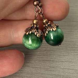 May include: A pair of earrings with green, spherical beads with a marbled effect. The beads are suspended from ornate, bronze-coloured metalwork. The earrings are shown against a neutral background.