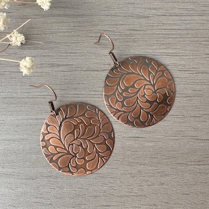 Op de afbeelding: Twee koperkleurige, ronde oorbellen met een bloemmotief. De oorbellen hebben een haakontwerp voor doorboorde oren. Het bloemmotief is in reliëf op het oppervlak van de oorbellen. De oorbellen worden gepresenteerd op een grijze houten ondergrond.