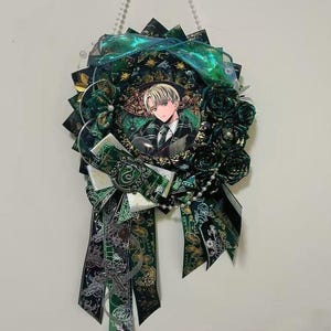 Könnte beinhalten: Ein dekoratives Abzeichen mit einem zentralen Porträt einer Person mit blonden Haaren, umgeben von grünen und schwarzen Bändern, Stoff und künstlichen Blumen. Das Abzeichen ist mit Perlen und einer Hängekette verziert.