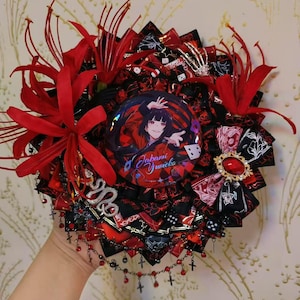 Könnte beinhalten: Dekoratives, mehrschichtiges Accessoire in Rot und Schwarz, mit einem zentralen Abzeichen mit Anime-Kunst. Das Design umfasst rote Blumen, Bänder, Würfel und ein Schlangenmotiv. Eine goldfarbene Brosche mit rotem Edelstein fügt Details hinzu.