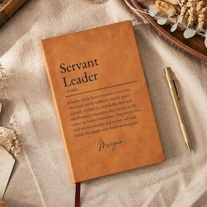 May include: A tan leather journal with the words "Servant Leader" and a definition printed on the cover. A gold pen rests beside the journal. The journal has a brown ribbon bookmark.