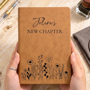 May include: A tan leather-like journal featuring the black text "Jolene's NEW CHAPTER" and a black line art illustration of various wildflowers at the bottom. The journal is held in place, surrounded by two pens, an inkwell, a pocket watch, and a key.