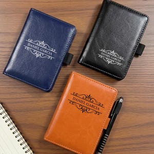 May include: Three rectangular passport holders in navy blue, black, and orange, each featuring a smooth finish and a decorative filigree design with the name DANIEL GARCIA printed in the centre. A black pen is attached to the orange holder.