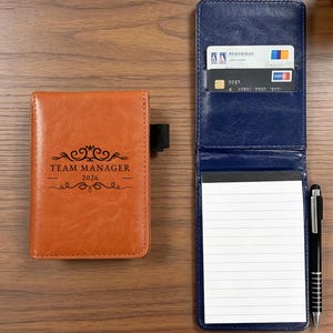 May include: A tan leather-like pocket notebook with the text TEAM MANAGER 2026 printed on the front cover, alongside an open navy blue leather-like notebook holder containing two credit cards and a lined notepad, with a black pen resting beside it.