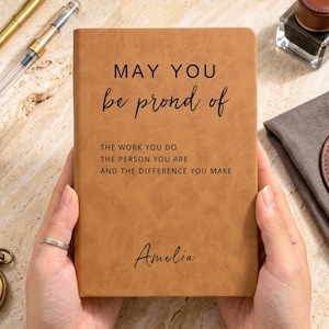 May include: A tan leather-textured notebook featuring the black printed text: MAY YOU be prond of THE WORK YOU DO THE PERSON YOU ARE AND THE DIFFERENCE YOU MAKE, with the name Amelia printed in a script font at the bottom of the cover.