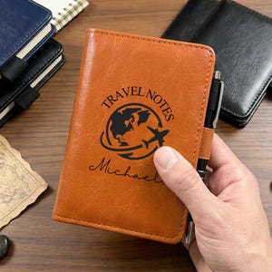 Puede incluir: Una funda para cuaderno de color canela con aspecto de cuero que presenta un gráfico negro de un globo terráqueo con un avión y el texto "TRAVEL NOTES" y "Michael" impreso en la parte frontal. Un bolígrafo negro se sujeta en una banda elástica.