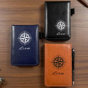 Può includere: Tre taccuini in pelle blu navy, neri e marrone chiaro, ciascuno con un design a rosa dei venti e il nome "Liam". Una penna è posizionata accanto al taccuino marrone chiaro. I taccuini sono su una superficie di legno.