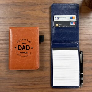 Puede incluir: Una funda de cuaderno de bolsillo color canela con el texto YOU ARE THE BEST DAD CHARLES HAPPY FATHER'S DAY grabado en la parte frontal. Al lado, un portacuadernos azul marino abierto contiene dos tarjetas de crédito, un bloc de notas y un bolígrafo negro.