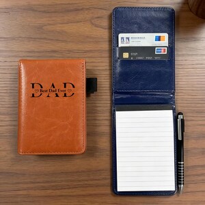 Könnte beinhalten: Ein braunes Leder-Notizbuch mit dem Text "DAD Best Dad Ever" liegt neben einem blauen Leder-Notizblock mit einem linierten Papierblock, Kreditkarten und einem Stift. Die Artikel befinden sich auf einer Holzoberfläche.