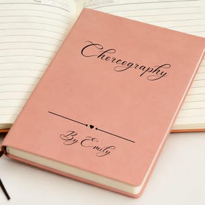 May include: A coral-coloured notebook with the word "Choreography" in elegant script. Below, a line with two hearts and the words "By Emily" are printed. The notebook is open to reveal lined pages.