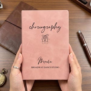 May include: A dusty pink notebook with a matte finish featuring the black text choreography, a line drawing of ballet slippers, the name Maeve, and the text BROADWAY DANCE STUDIO printed on the front cover in a clean, sans-serif font.