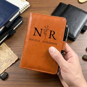 May include: A tan leather-like notebook cover featuring the black printed initials N R with a floral stem between them and the name NICOLE ROBINSON below. A black pen is held in the elastic loop on the side of the cover, which has a textured finish.