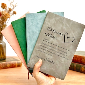 Puede incluir: Un conjunto de cuatro cuadernos rectangulares con cubiertas texturizadas en gris, azul claro, verde y rosa claro. El cuaderno gris presenta el texto Difference Maker noun, un símbolo de corazón, una definición y el nombre Emily en negro.